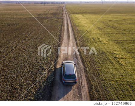 Aerial view of car driving by straight ground road through green fields on sunny blue sky copy space background. Drone photography. Aerial view of car driving by straight ground road through green fields on sunny blue sky copy space background. Drone photography. 132749969