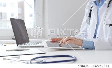 Female doctor wearing lab coat and stethoscope using digital tablet with laptop and medical records on desk in medical office, showcasing modern healthcare technology. Medicine concept 132752854
