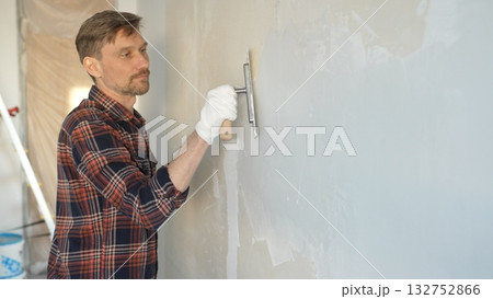 Male construction worker wearing gloves is carefully applying plaster to a wall using a hand tool, contributing to a home renovation or construction project 132752866
