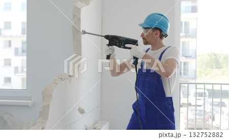 Male construction worker demolishing concrete wall with rotary hammer, wearing work overalls and blue hardhat is generating dust and debris during home renovation work, portrait horizontal view 132752882