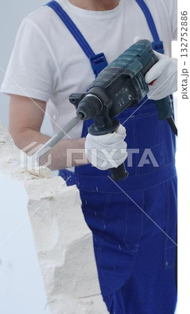 Construction worker demolishing white wall with rotary hammer drill, wearing blue overalls and protective white gloves, generating dust during renovation project, closeup view 132752886
