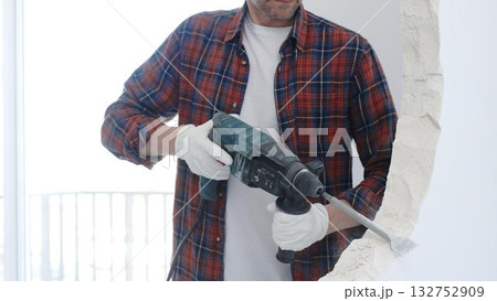 Male construction worker wearing red checkered shirt and protective white gloves is demolishing white wall with rotary hammer drill, closeup view Male construction worker wearing red checkered shirt and protective white gloves is demolishing white wall with rotary hammer drill, closeup view 132752909