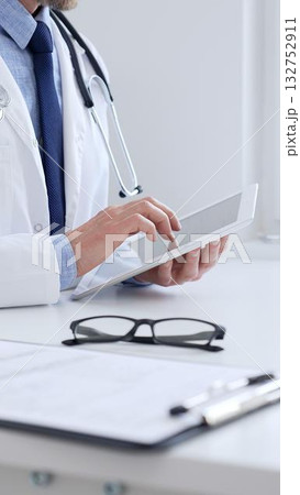 Adult male doctor wearing lab coat and stethoscope using digital tablet while sitting at desk in medical office, close up and vertical view. Medicine, health care concept Adult male doctor wearing lab coat and stethoscope using digital tablet while sitting at desk in medical office, close up and vertical view. Medicine, health care concept 132752911