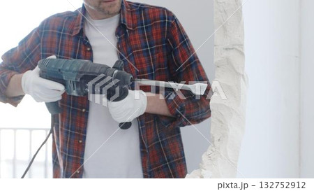 Male construction worker wearing red checkered shirt and protective white gloves is demolishing white wall with rotary hammer drill, closeup view 132752912