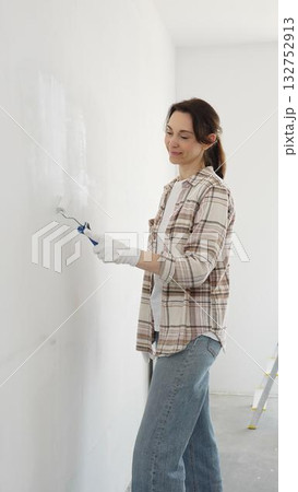 Woman wearing protective gloves rolling white paint onto wall, updating living space with fresh color and personal home renovation effort 132752913