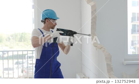 Male construction worker demolishing concrete wall with rotary hammer, wearing work overalls and blue hardhat is generating dust and debris during home renovation work, portrait horizontal view Male construction worker demolishing concrete wall with rotary hammer, wearing work overalls and blue hardhat is generating dust and debris during home renovation work, portrait horizontal view 132752948