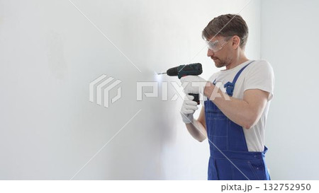 Skilled worker drilling hole in white wall, wearing protective gear during home improvement project with cordless power tool 132752950