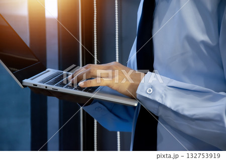A businessman stands near a bright window and is using a laptop. 132753919