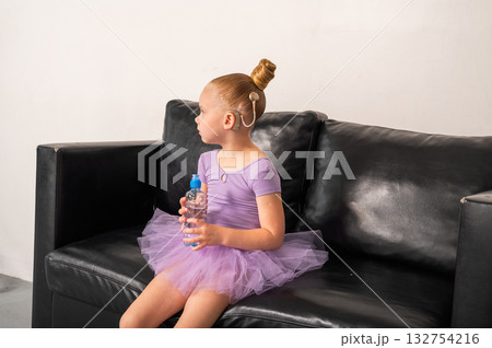 Girl ballerina with cochlear implant sitting on black sofa holding bottle of water. Inclusion, childhood recovery, and empowerment through hearing and rest. 132754216
