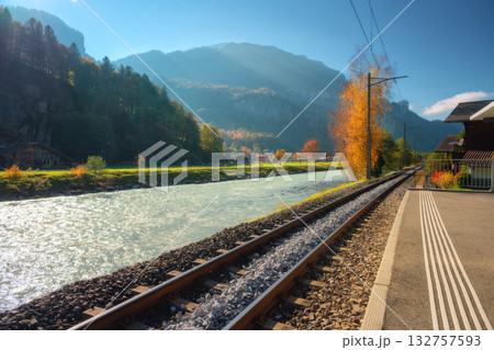 Railway tracks along a river, autumn trees and alpine mountains 132757593