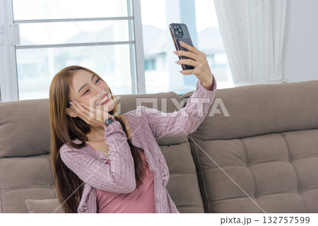 Young woman happy at home enjoys people activity, taking charming selfie with mobile phone, smiling cheerfully from sofa in bright living room, sharing joyful moment. 132757599