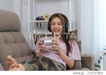 Young woman, happy people at home enjoy gaming activity, wearing headphones, relaxing on sofa with controller, playing video games, leisure time spent indoors. 132757601