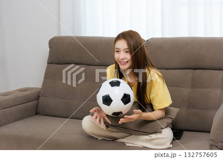 Young happy woman at home enjoying sport activity, sitting comfortably on cozy sofa, holding soccer ball, eagerly anticipating game with bright smile, showing cheerful spirit. 132757605