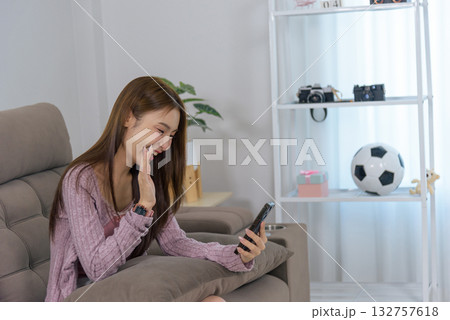 Young happy woman enjoys home activity, communicating with phone with people, waving hand, smiling wide, sharing laughter, leisurely moment relaxing on sofa, casual style, modern living room. 132757618