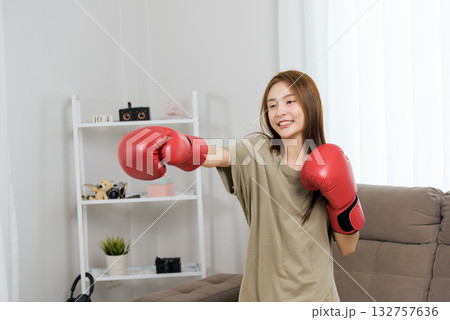 Young happy woman performs energetic activity at home, engaging in boxing fitness, punching with red gloves, demonstrating strength and healthy lifestyle dedication indoors. 132757636