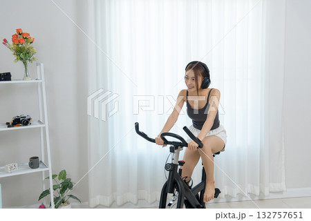 Young woman happy at home enjoying activity, people pursuing fitness, exercising on stationary bike, listening with headphones, healthy lifestyle, promoting wellbeing. 132757651