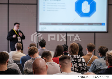 Presenter delivers talk to engaged crowd in conference room with large projection screen Presenter delivers talk to engaged crowd in conference room with large projection screen 132758866