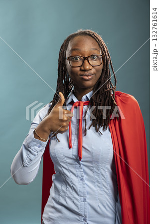African american female superhero in blue shirt and red cape gives a thumbs up against isolated background. Her confident gesture radiates approval, positivity and professional charm. African american female superhero in blue shirt and red cape gives a thumbs up against isolated background. Her confident gesture radiates approval, positivity and professional charm. 132761614