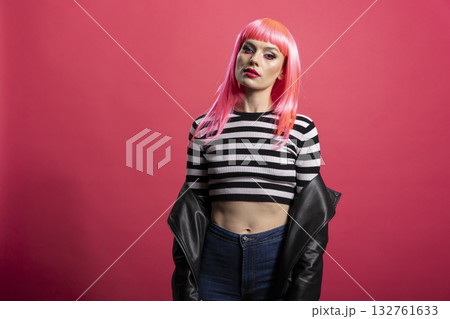 Gorgeous white woman with vibrant hairstyle and striped crop top stands poised and looks at camera. Beautiful young lady with black jacket, expresses calm confidence in pink background studio. Gorgeous white woman with vibrant hairstyle and striped crop top stands poised and looks at camera. Beautiful young lady with black jacket, expresses calm confidence in pink background studio. 132761633