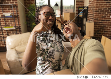 Cheerful boyfriend and girlfriend take playful selfie together, holding keys and cardboard box after moving into new home. Man holds phone, making goofy face and enjoying moment with partner. Cheerful boyfriend and girlfriend take playful selfie together, holding keys and cardboard box after moving into new home. Man holds phone, making goofy face and enjoying moment with partner. 132761639