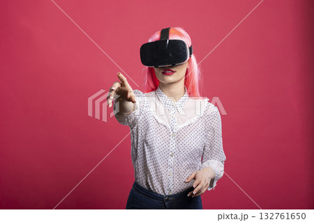 Young woman with pink wig wearing VR headset, points finger toward camera during interaction with immersive 3D environment. Curious caucasian female explores futuristic digital world. Young woman with pink wig wearing VR headset, points finger toward camera during interaction with immersive 3D environment. Curious caucasian female explores futuristic digital world. 132761650