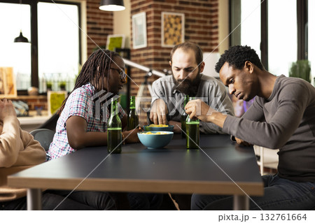 Young male and female adults sit around table with beer and chips, looking bored in cozy room. Group of friends relax together in brick walled room, sharing snacks and drinks, feeling unoccupied. Young male and female adults sit around table with beer and chips, looking bored in cozy room. Group of friends relax together in brick walled room, sharing snacks and drinks, feeling unoccupied. 132761664