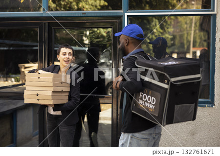 Black courier handing a heavy pile of pizza boxes to a young woman, express takeout service. Male employee brings takeaway meal from a fast food restaurant in an urban setting. 132761671