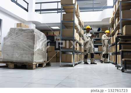 Black female worker handling orders and supply package shipments, managing goods in a depot warehouse. Distribution products arranged on racks ready for delivery, import export. 132761723