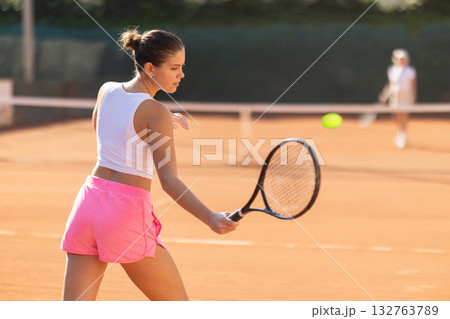Back view of sporty young girl spending time playing tennis with racket and ball on tennis court 132763789