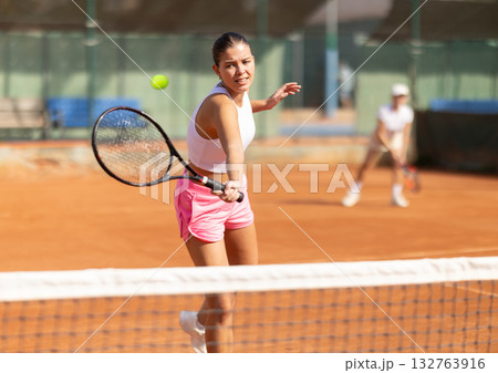 Active young girl hits tennis ball with racket in front of players in court. Two sportive women enjoying padel tennis Active young girl hits tennis ball with racket in front of players in court. Two sportive women enjoying padel tennis 132763916