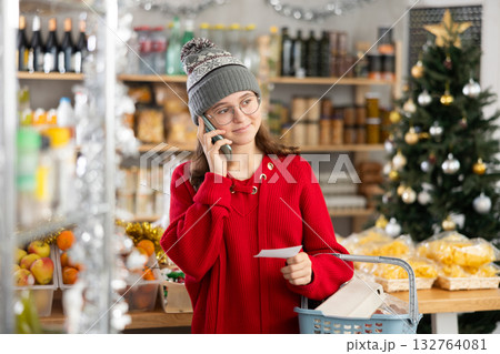 She holds paper note in hands, asks mother questions on phone while shopping in grocery 132764081