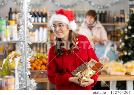 Positive girl in festive hat choosing sweets for New Year's holiday in supermarket with mother choosing food on shelves in Positive girl in festive hat choosing sweets for New Year's holiday in supermarket with mother choosing food on shelves in 132764267