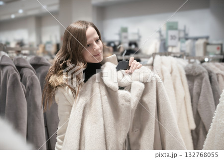 Young woman tries on cozy white fur coat in fashion boutique for festive autumn winter shopping on Christmas Eve Young woman tries on cozy white fur coat in fashion boutique for festive autumn winter shopping on Christmas Eve 132768055