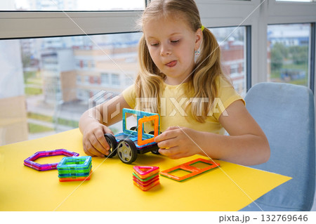 The photo shows a little girl creatively playing with magnetic building blocks, constructing various designs against a solid yellow background for contrast. The photo shows a little girl creatively playing with magnetic building blocks, constructing various designs against a solid yellow background for contrast. 132769646