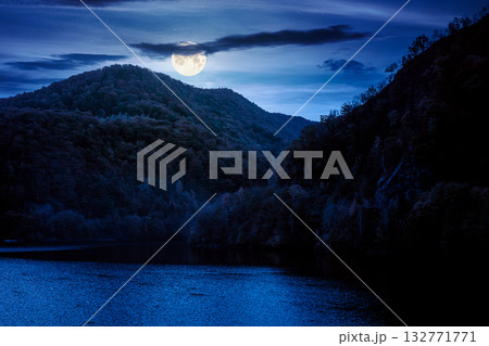mountainous landscape with lake in autumn at night. forest on hills near the shore of river under dark sky in full moon light. beautiful view of stunning place 132771771