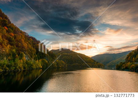 mountainous landscape with lake tarnita in autumn at dusk. forest on hills near the shore of somes river under orange sky. beautiful view in apuseni nature park romania, europe. stunning place mountainous landscape with lake tarnita in autumn at dusk. forest on hills near the shore of somes river under orange sky. beautiful view in apuseni nature park romania, europe. stunning place 132771773