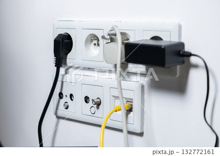 Busy wall outlet with multiple electrical plugs, network cables, and various charging adapters connected, showcasing essential modern home technology connectivity and digital energy needs 132772161