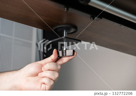 Person's hand tightening a sturdy desk clamp, installing an accessory underneath a wooden table for an organized home office workspace setup 132772206