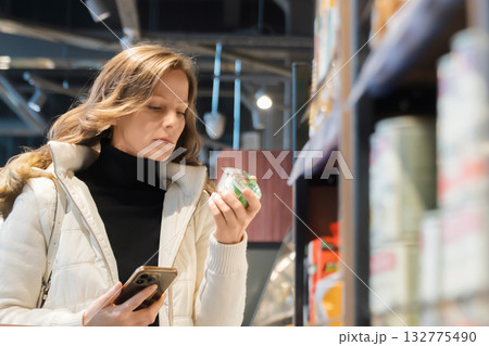 A blonde woman studies the label of a jar of yogurt, focusing on its ingredients and shelf life, while shopping for healthy food in a grocery store. 132775490