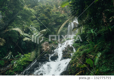 A beautiful waterfall cascades down from a rocky cliff amidst lush greenery and mist at the base of the waterfall. 132775755