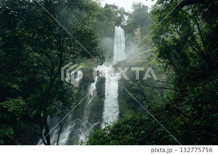A beautiful waterfall cascades down from a rocky cliff amidst lush greenery and mist at the base of the waterfall. A beautiful waterfall cascades down from a rocky cliff amidst lush greenery and mist at the base of the waterfall. 132775756