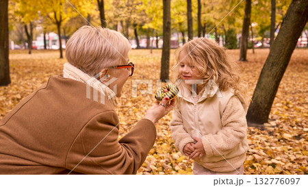 Grandmother offering donut to little girl in yellow leaves park Grandmother offering donut to little girl in yellow leaves park 132776097