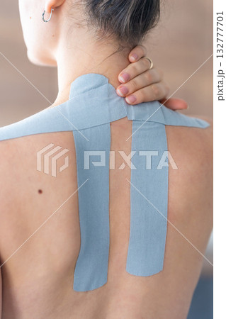 Close up view of young woman with kinesiology tape applied to her neck and shoulder holding her neck with her hand. Physical therapy and muscle recovery through modern rehabilitative care Close up view of young woman with kinesiology tape applied to her neck and shoulder holding her neck with her hand. Physical therapy and muscle recovery through modern rehabilitative care 132777701