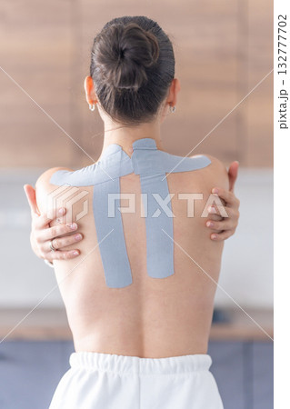 Young woman with kinesiology tape on her back and shoulders, embracing herself with both hands. Physical therapy, body awareness and emotional self-care through healing touch Young woman with kinesiology tape on her back and shoulders, embracing herself with both hands. Physical therapy, body awareness and emotional self-care through healing touch 132777702