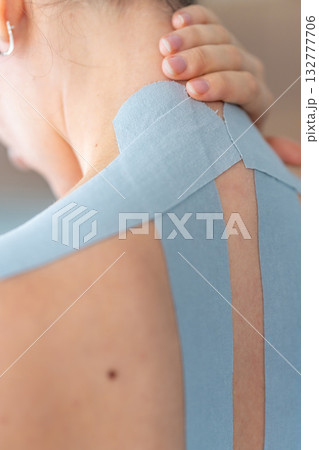 Close up view of young woman with kinesiology tape applied to her neck and shoulder holding her neck with her hand. Physical therapy and muscle recovery through modern rehabilitative care Close up view of young woman with kinesiology tape applied to her neck and shoulder holding her neck with her hand. Physical therapy and muscle recovery through modern rehabilitative care 132777706