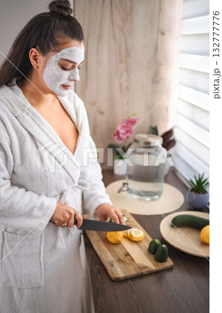 Young woman wearing bathrobe and face mask cutting lemon in the kitchen Young woman wearing bathrobe and face mask cutting lemon in the kitchen 132777776