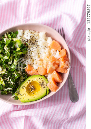 Fresh breakfast bowl featuring salmon, sliced avocado, leafy greens, and rice served on a pink striped tablecloth. Perfect for a nutritious start to the day 132779897
