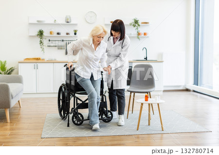 A female doctor helps an older woman stand up from her wheelchair in a well-lit room, promoting mobility and care. A female doctor helps an older woman stand up from her wheelchair in a well-lit room, promoting mobility and care. 132780714