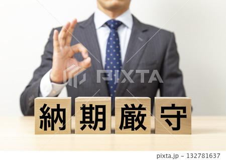 「納期厳守」と書かれたブロックとOKポーズをするビジネスマン 「納期厳守」と書かれたブロックとOKポーズをするビジネスマン 132781637