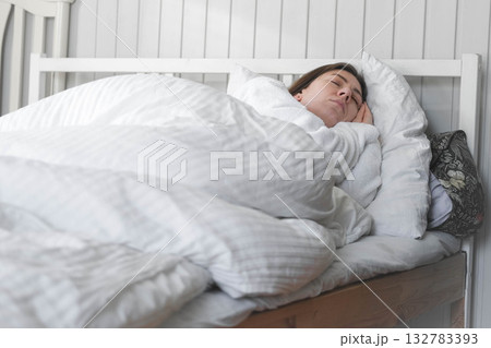 A woman is sleeping peacefully in her bed with a white pillow and a blanket. A beautiful young happy lady is sleeping peacefully on a cozy bed in the morning light at home, in a relaxed atmosphere A woman is sleeping peacefully in her bed with a white pillow and a blanket. A beautiful young happy lady is sleeping peacefully on a cozy bed in the morning light at home, in a relaxed atmosphere 132783393